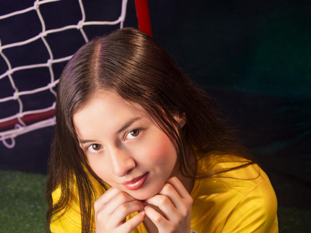VioletaNova, com cabelo castanho, camisa amarela e mãos perto do rosto, olhando para cima com um leve sorriso, mostrando seus lábios rosados e pele macia. Rede de futebol ao fundo..