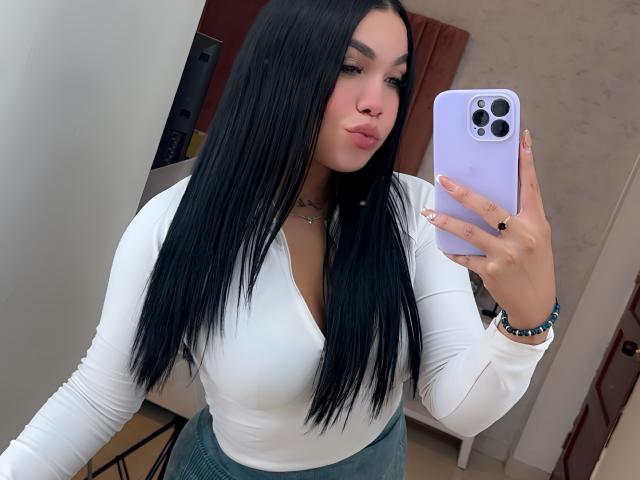 MiaDenver with long black hair, white long-sleeve top, and green skirt, holding purple phone, pouting lips, showing small tits, wearing bracelet and ring, in selfie.