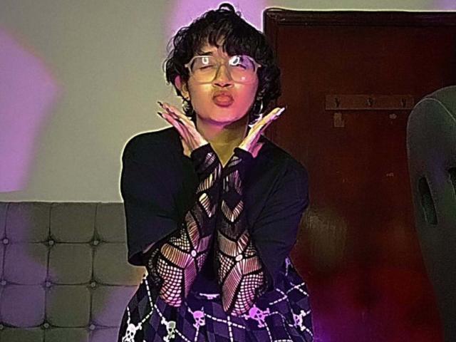 DaryMoon with short black curly hair, glasses, pouty lips, and hands up, wearing a black top and fishnet sleeves, standing against a wooden door.