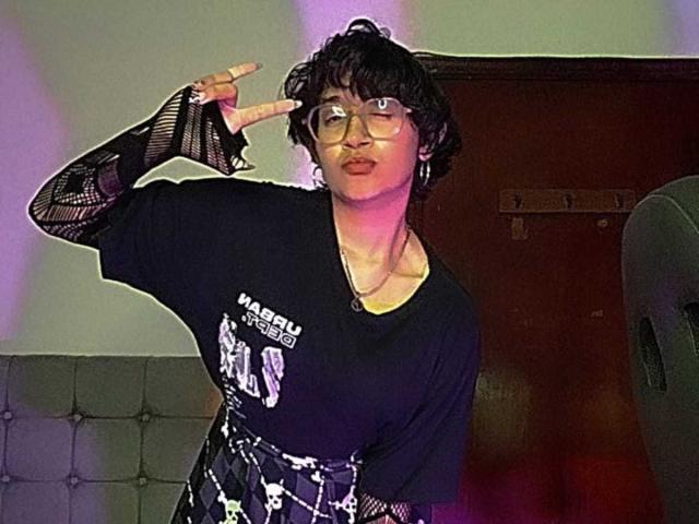 DaryMoon with curly black hair, glasses, and black mesh sleeves, making a rock 'n' roll gesture, wearing a black shirt and patterned skirt, standing in a dimly lit room.