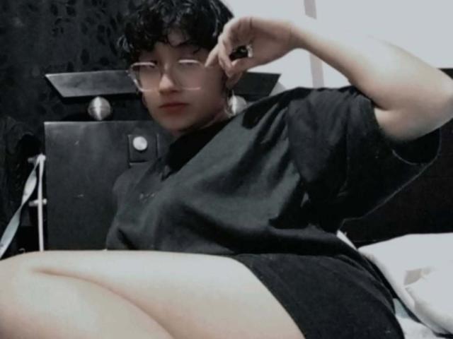 DaryMoon, with short curly black hair and glasses, sits on a bed in a black shirt and shorts, adjusting his glasses with a relaxed, slightly confident expression.