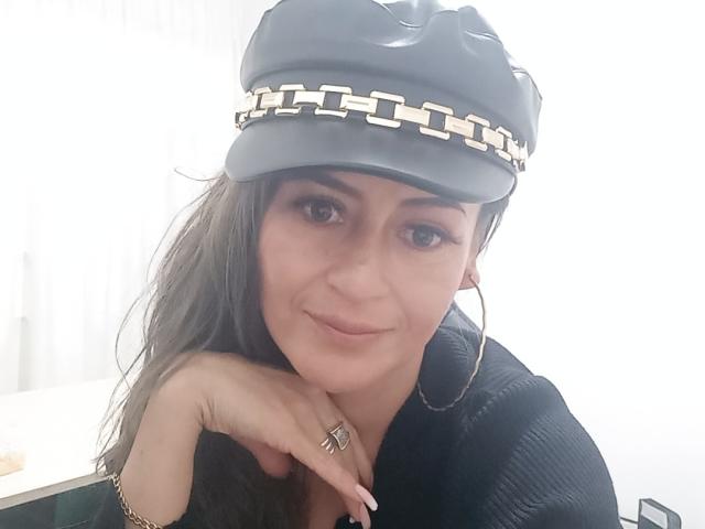 Maghentaa, con cabello largo y castaño, lleva un gorro de cuero negro con una cadena de plata, una blusa negra y aros grandes, sonríe suavemente, con el mentón apoyado en la mano, mostrando un anillo..