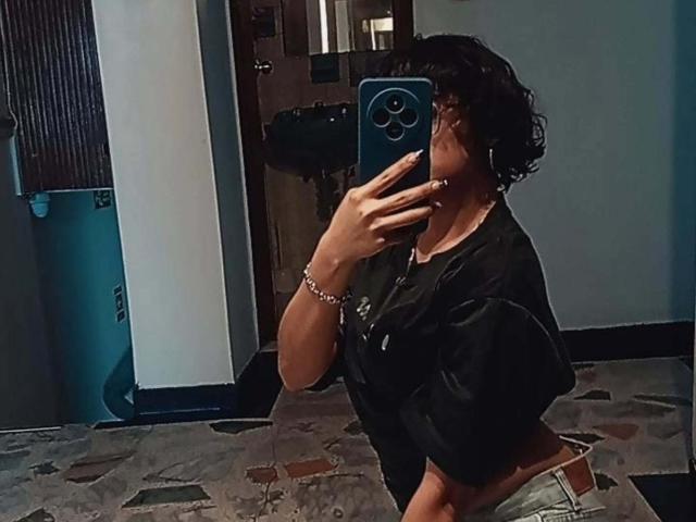 DaryMoon with curly black hair, wearing a black shirt and beige pants, taking a mirror selfie with a blue phone, showing a slight view of her ass.