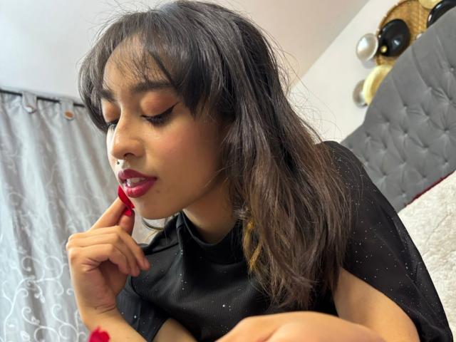 CamiVargas with dark, wavy hair, red lipstick, and black dress, biting red fingernail, eyes closed, soft makeup, subtle blush, cute, attractive, bedroom, tufted headboard, white curtains.