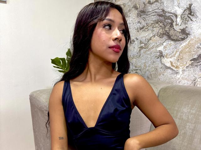 GabyVegaa, con cabello negro largo, lápiz labial rosa oscuro y un vestido azul marino intenso, que muestra pequeños senos y un tatuaje en su brazo derecho, mirando hacia un lado..