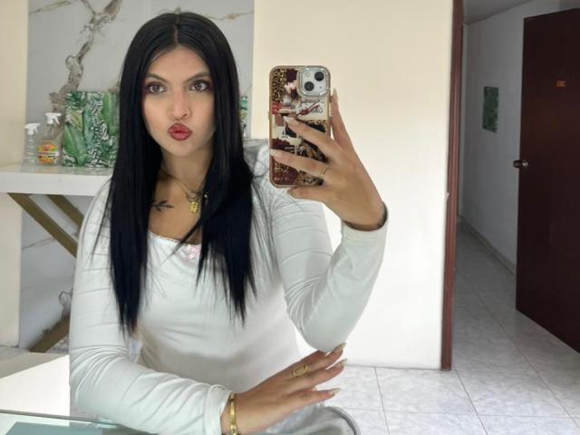 NyraVanel, avec ses cheveux noirs, son rouge à lèvres rouge, sa chemise blanche à manches longues, tenant un téléphone avec une coque à motifs serpent, se prend en selfie devant un miroir, montrant de petites seins et des fesses, dans une pièce carrelée..
