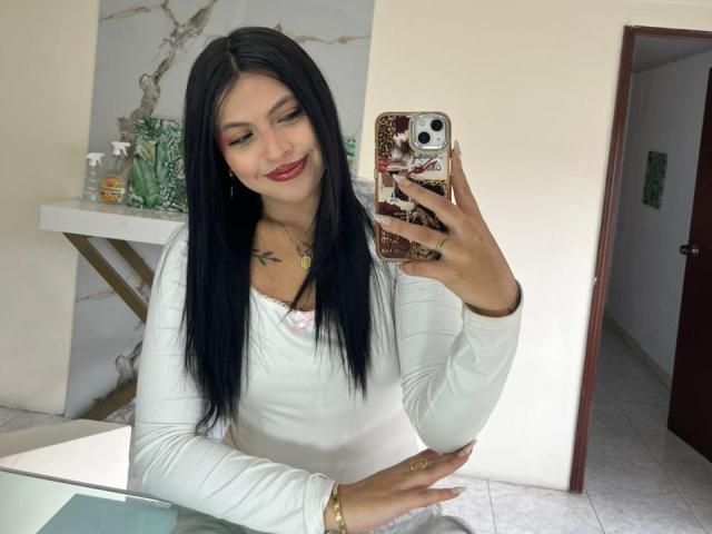 NyraVanel with long black hair, white long-sleeve top, red lipstick, taking a mirror selfie with a phone, showing a tattoo on her chest, smiling. Boobs visible under top.