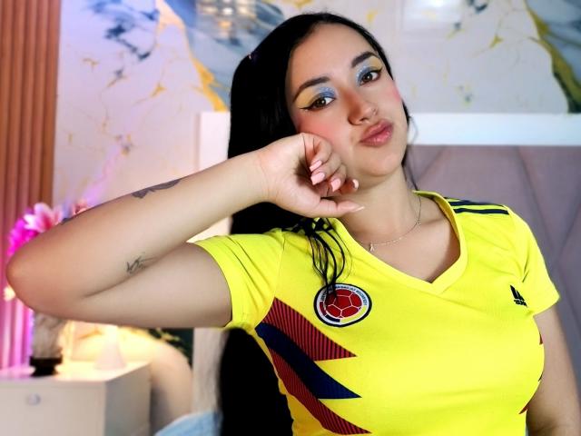 MiiaRousse in a yellow soccer jersey, pouting with a hand on her cheek, blue eye makeup, black hair, small tattoo on her arm, and a cute, sexy face.