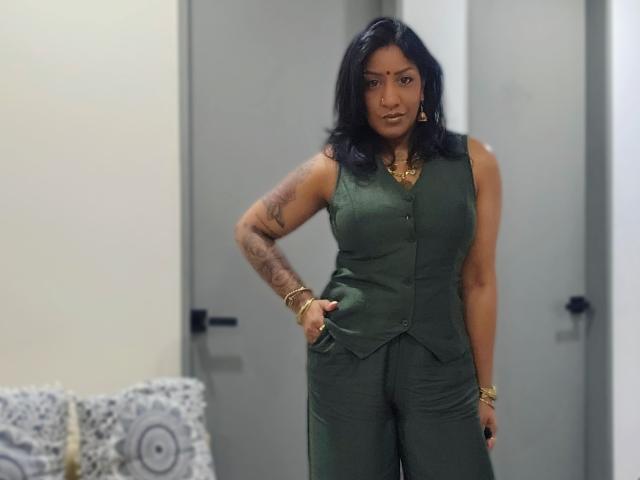 Nahirr ha i capelli scuri, un braccio tatuato e indossa un gilet verde senza maniche con dei pantaloni, gioielli dorati e ha un'espressione seria, rimanendo in piedi con una mano in tasca..