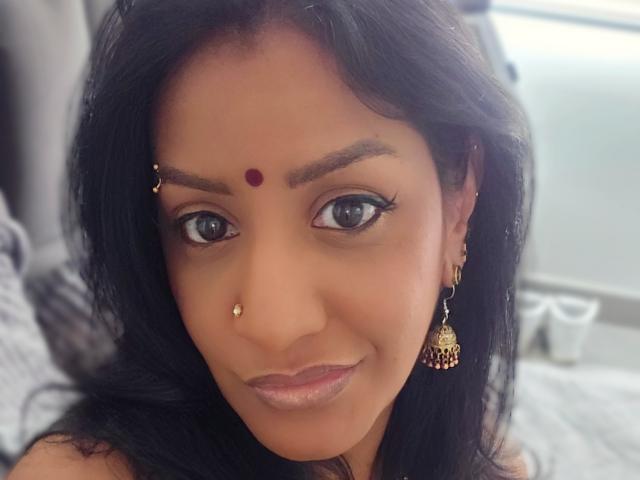 Nahirr has dark hair, a red bindi on her forehead, a gold nose ring, and a gold earring with a intricate design, looking at the camera with a smile.