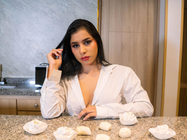 MelodyRoss, con i capelli neri, una camicia bianca e un profondo scollo, è in piedi in una cucina, toccandosi i capelli, con dell'impasto per dolci bianco su un piano di lavoro in granito..