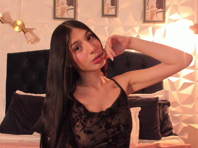 Sophie Beaumont, con cabello negro largo, vistiendo una blusa negra transparente que deja ver pequeños senos, tiene un tatuaje en la clavícula izquierda, con un dedo cerca de la boca, un cabecero de terciopelo negro y una luz cálida y suave..