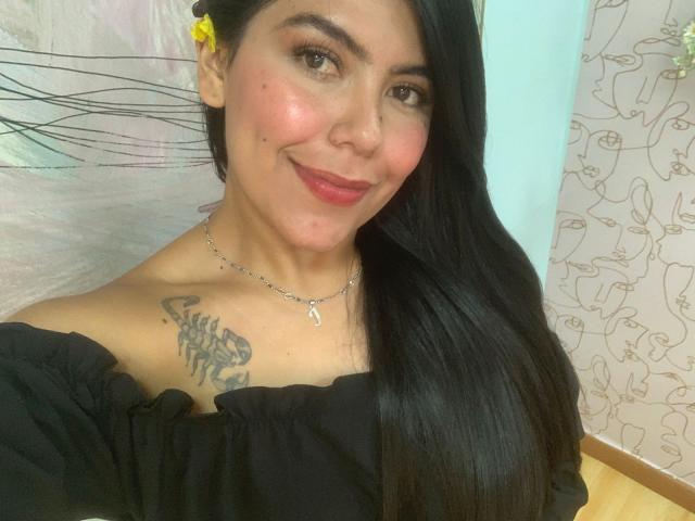 CatalinaReiyna has black hair, red lipstick, and a cute smile. She's wearing an off-shoulder black top, showing a tattoo on her right shoulder. She's wearing a yellow flower.