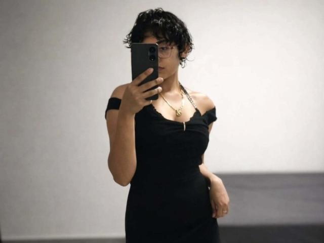 DaryMoon with curly black hair, glasses, black off-shoulder dress, necklace, holding phone in mirror selfie, showing tits and arms, in a minimalist room.