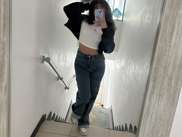 SofiaPassionne in a mirror selfie, wearing a black jacket, white crop top, and high-waisted blue jeans, with curly hair, holding a phone, showing her flat midriff and ass.