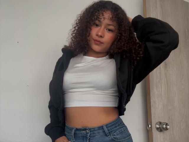 SofiaPassionne with curly brown hair, wearing a black zip-up jacket, white crop top, and blue jeans, hand in jacket, standing against a door. Her ass is visible.