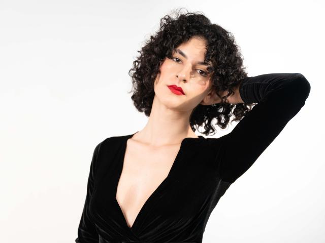 RoxyGraff, avec ses cheveux noirs bouclés, portant une robe en velours noir profond qui dévoile sa poitrine, avec du rouge à lèvres et une main dans ses cheveux, regardant légèrement sur le côté..