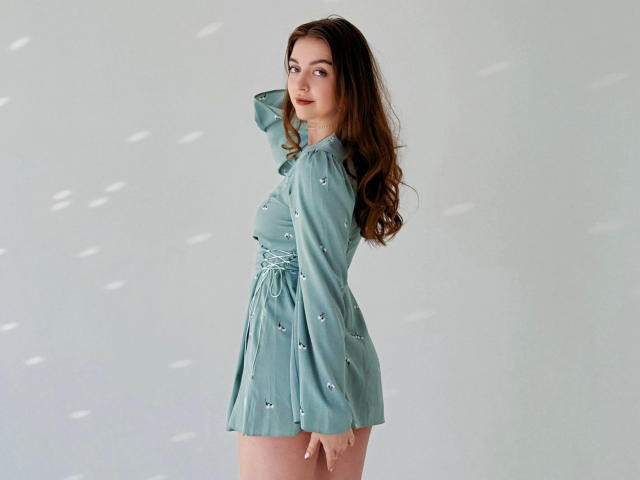 MaryJoy-hot with long brown hair, wearing a teal dress with small white patterns, standing sideways, hand on head, looking at camera, soft light, subtle shadows on wall.