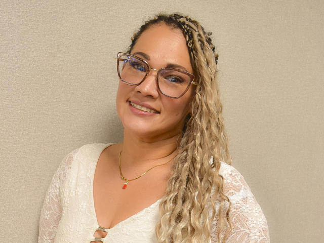 Vianca Castellanos, avec des tresses blondes bouclées, portant des lunettes, un haut en dentelle blanche, un collier en or avec un pendentif rouge, souriant à la caméra, montrant sa poitrine et un joli visage..