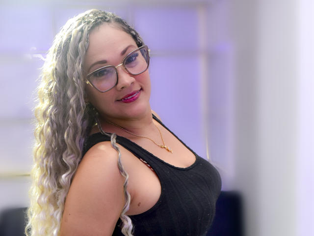 Vianca Castellanos, met krullend grijs haar, een bril, roze lippenstift, een zwart tanktopje, een gouden ketting, lacht naar de camera en laat haar borsten zien. Zacht, binnenlicht..