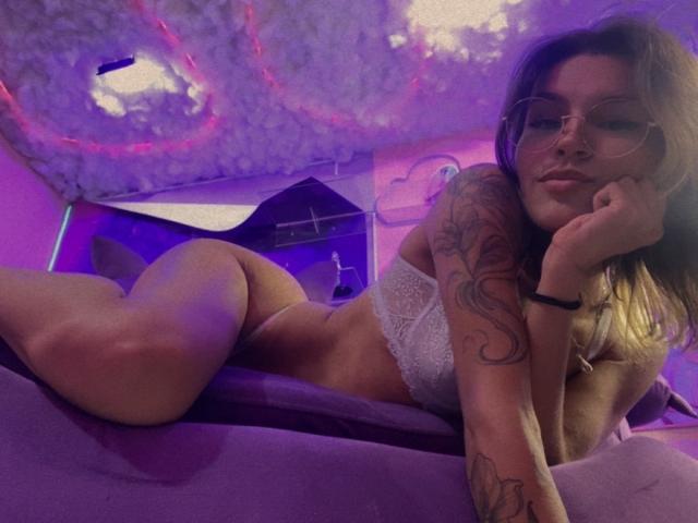 LenaBranch lies on a purple bed, wearing white lace panties and a matching bra, with glasses and tattoos on her arm, her ass raised, her face resting on her hand.