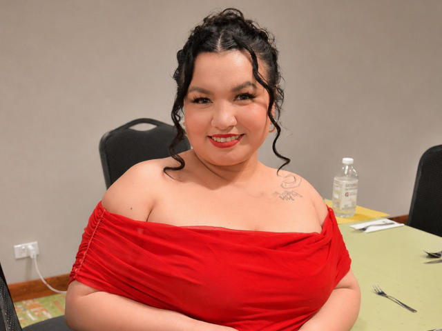 LillyMayer, chubby Asian woman with curly black hair, red off-shoulder dress, small tattoo on right shoulder, smiling, arms crossed, table with water bottle in background.