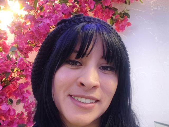 IssisDiosa heeft zwart haar, een pony en draagt een gebreide muts. Ze glimlacht met witte tanden, heeft een lichtroze huid en bruine ogen, en is omringd door felroze bougainvillea-bloemen..