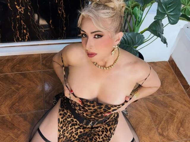 MisseScarlet, con cabello rubio, vestida con un vestido de estampado de leopardo, mostrando sus grandes senos, con las manos sobre el pecho, un collar dorado, lápiz labial rojo y tirantes negros hasta los muslos, sentada en un suelo de madera..