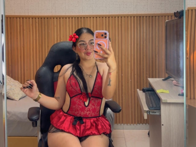 LunaLille draagt rode kant lingerie, met haar borsten zichtbaar in een spiegel-selfie, draagt een bril, heeft een rode bloem in haar haar, staat voor een zwarte stoel, een bureau met een toetsenbord, houdt een telefoon vast, en er is een houten muur op de achtergrond..