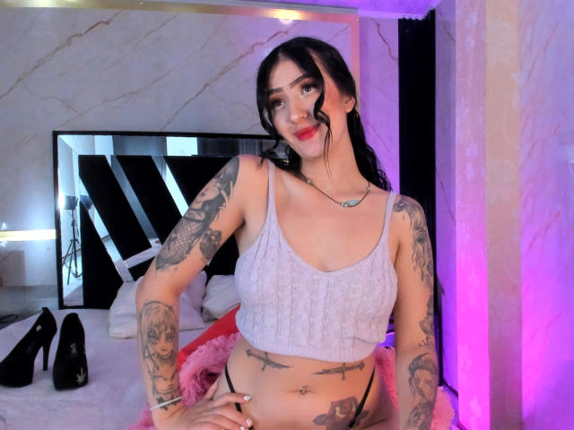 TifanyDiaz ha dei tatuaggi, indossa una canotta grigia che mostra il seno e l'ombelico, con biancheria intima rosa, tacchi alti neri e una collana, in una stanza molto luminosa..