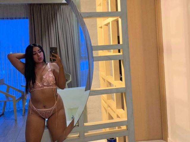 BambyVall está em pé, vestindo lingerie de renda rosa, mostrando seus seios e vulva, segurando um telefone, com cabelos longos e pretos, em frente a um espelho em um quarto moderno..
