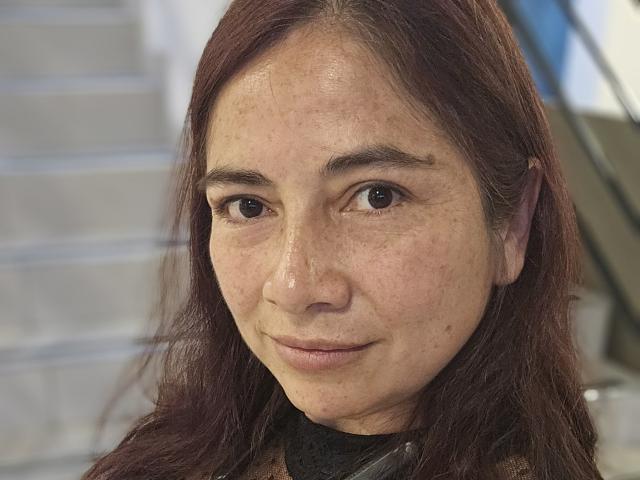 AlessiaRed ha un viso carino, con occhi marroni, leggere lentiggini e lunghi capelli castano-rossicci. Indossa una collana nera e ha un sorriso leggero e rilassato..