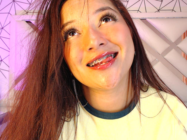 NicoleMartins tiene el pelo largo y castaño, ojos marrones y una gran sonrisa con brackets metálicos, lleva una camisa blanca con cuello verde oscuro, y muestra sus dientes brillantes..