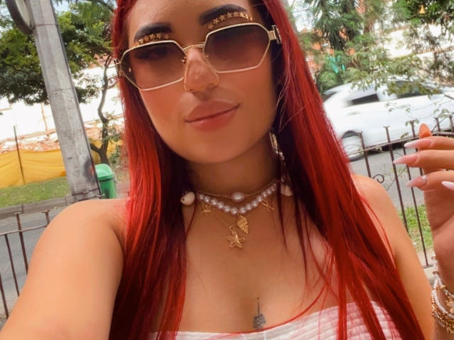 MeganCherry: Een vrouw met rood haar, die gouden zonnebrillen met oogjes, een parelketting en een geruit top draagt. Ze laat een kleine tatoeage op haar borst zien en heeft een lichte glimlach..