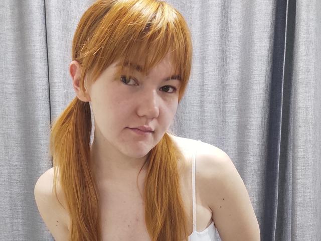 SheritaChieng, com rabos de cavalo castanho claro, camiseta branca, seios pequenos, pele clara, sorriso discreto, olhando para a câmera, fundo com cortina cinza..