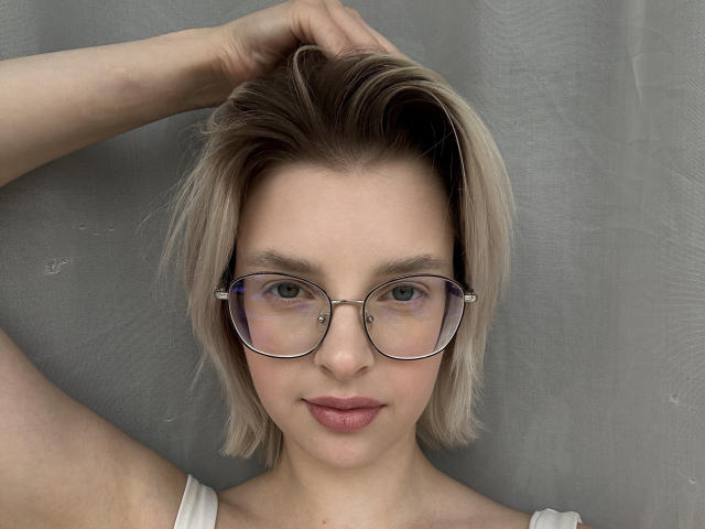 BrandeKocur ha i capelli biondi e corti, indossa occhiali rotondi con un piercing al naso e ha un sorriso discreto. Sta toccando la testa con un braccio e indossa una maglietta bianca..
