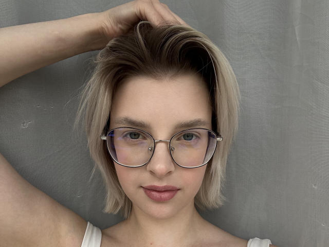 BrandeKocur mit kurzen, blonden Haaren, Brille und rosafarbenen Lippen, mit erhobenen Armen, die ein Nasenpiercing zeigt, trägt ein weißes Oberteil und hat ein süßes Gesicht mit einem leichten Lächeln..