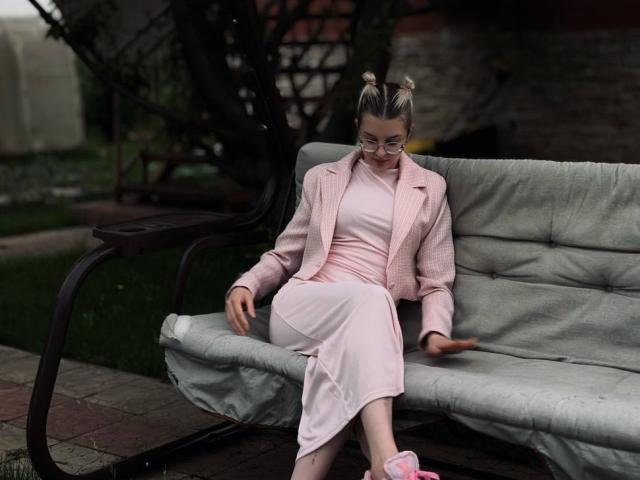 BrandeKocur mit blonden Zöpfen, Brille, einem pinken Outfit und einer Jacke, sitzt auf einer grünen Metallbank, die Beine gekreuzt, trägt pinke Slipper und blickt nach unten..
