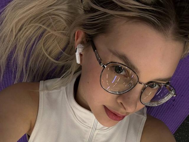 BrandeKocur, con i capelli biondi, indossa una canotta bianca, occhiali trasparenti, auricolari bianchi e ha le labbra rosa, è sdraiata su una superficie viola e guarda verso l'alto. Il seno non è visibile..