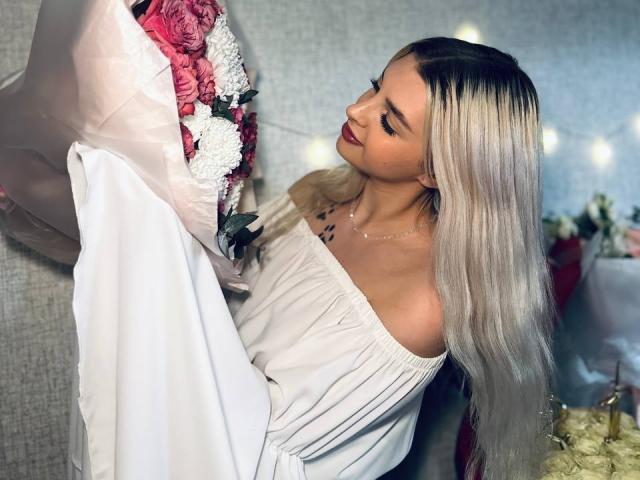 BrandeKocur mit langem, glattem, blondem Haar, die einen Strauß aus rosa und weißen Blumen hält, ein weißes Kleid mit einem Schulterfrei-Schnitt trägt und nach unten schaut, in einem sanft beleuchteten Raum..