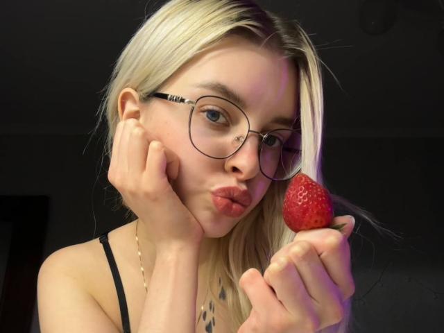 BrandeKocur mit blonden Haaren, Brille und Nasenring, zieht die Lippen zusammen, während sie eine rote Erdbeere an ihren Mund hält, ein schwarzer Träger ist auf ihrer Schulter sichtbar..