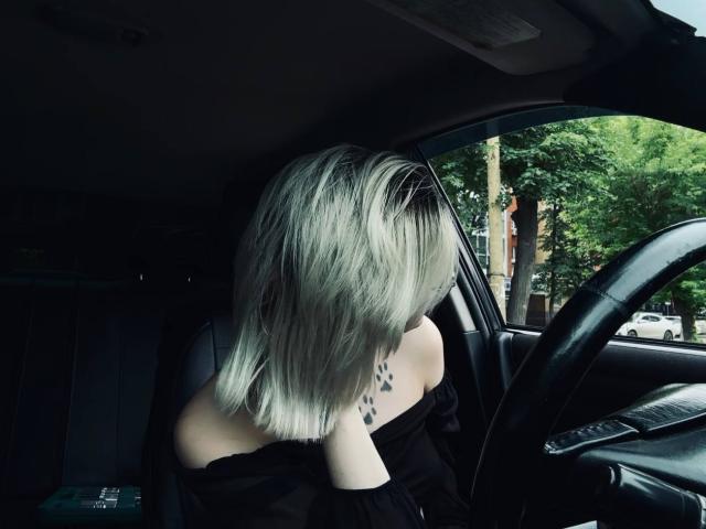 BrandeKocurs platinblondes Haar bedeckt ihren Hintern, während sie in einem Auto sitzt und ein schwarzes Oberteil mit einem Schulterauschnitt trägt, mit einem schwarzen Tattoo auf ihrem linken Schulterblatt..