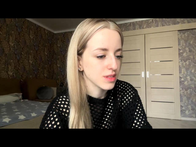 Maximina Mayhood mit blonden Haaren, heller Haut und rosafarbenem Lippenstift, trägt ein schwarzes Oberteil mit Punkten und blickt leicht nach unten. Ihr Hintern ist nicht sichtbar. Sie befindet sich in einem Schlafzimmer..