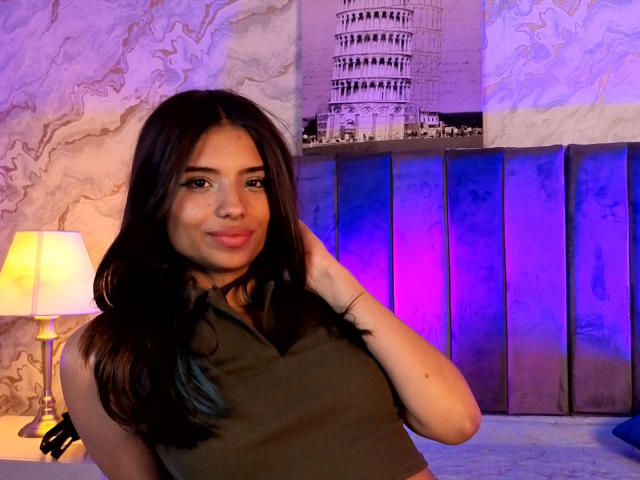 MadyPalmer with dark hair, wearing a green sleeveless top, smiling, hand in hair, purple and blue lighting, lamp on left, marble wallpaper, and a tall clock in background.