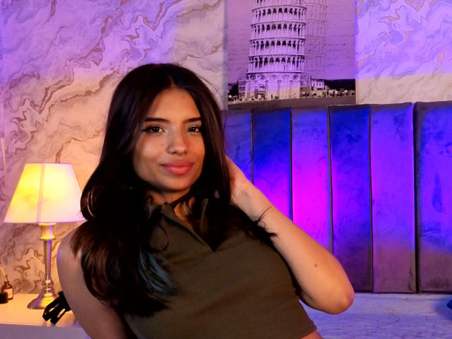 MadyPalmer has dark brown hair, medium tits, and a cute smile, wearing a green sleeveless top, with a purple and blue-lit background and a white lamp on the left.
