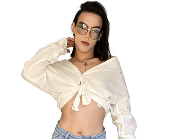 Melissa, con gli occhiali, indossa una camicia bianca annodata che lascia intravedere il seno, jeans blu e una collana, e guarda leggermente di lato con una mano vicino alla testa..