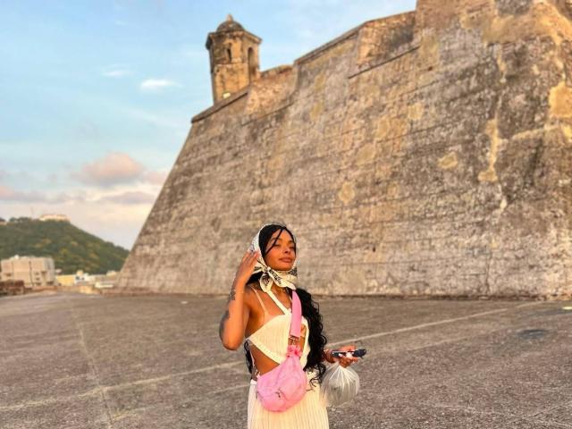 EmmaEbony stands in front of a massive stone wall, wearing a white dress with pink and black accents, black hair in a bun, holding a phone, and looking at camera.