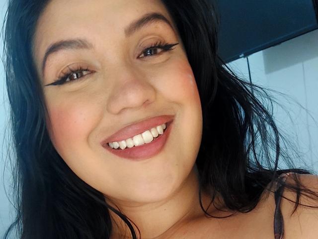 Laura Richis, con cabello negro, ojos marrones y largas pestañas, sonriendo mostrando sus dientes, con un lápiz labial rosado, y mostrando ligeramente sus senos en la parte superior de la imagen..