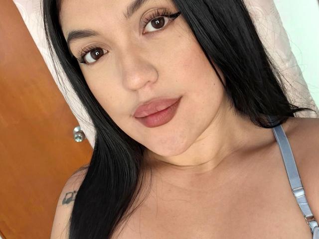 LauraRichis a les cheveux noirs, les yeux marrons et des lèvres pulpeuses avec un rouge à lèvres brun rosé. Elle porte une bretelle grise, a un tatouage sur l'épaule gauche et une serviette blanche..