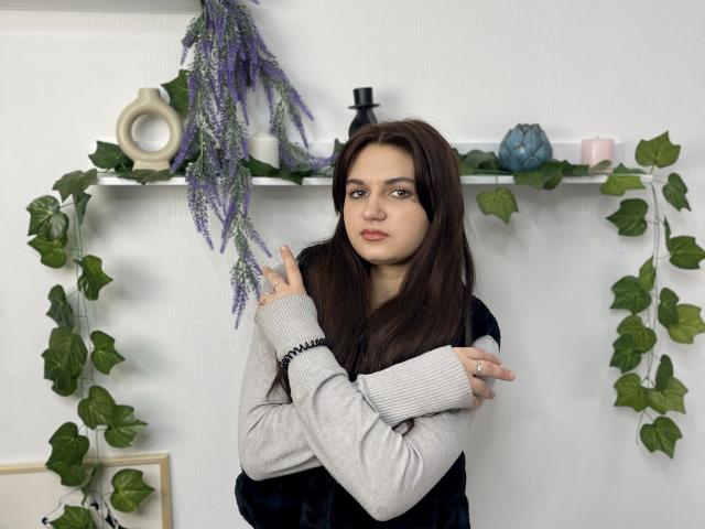 RamonaOsazuwa with long brown hair, light skin, and small tits, wearing a gray and black sweater, holding purple lavender, green vines, and white shelf with candles in the background.